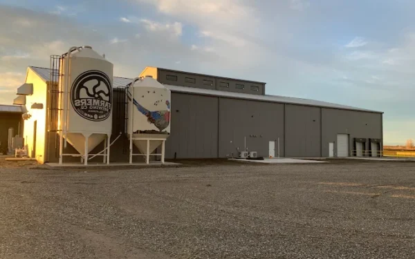 A large gray industrial building with two white silos labeled “Farmers Brewing Co.” stands on a gravel lot under partly cloudy skies.