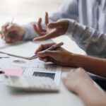Two people discuss financial charts and graphs at a desk, using pens and a calculator, with hands visible and documents spread out.