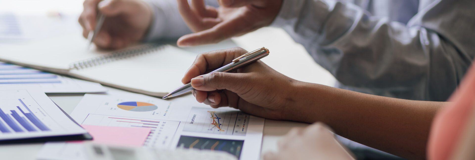 Two people discuss financial charts and graphs at a desk, using pens and a calculator, with hands visible and documents spread out.