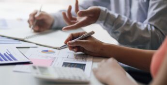 Two people discuss financial charts and graphs at a desk, using pens and a calculator, with hands visible and documents spread out.