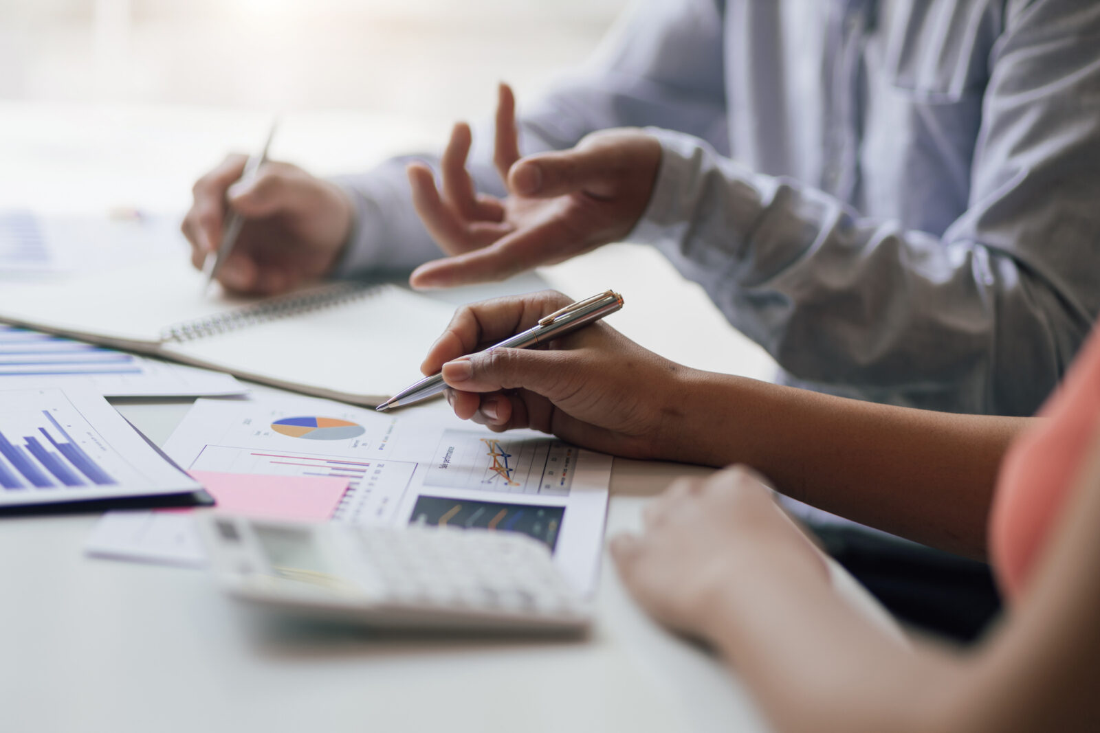 Two people discuss financial charts and graphs at a desk, using pens and a calculator, with hands visible and documents spread out.
