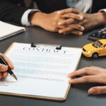 Two people sit at a desk with a contract, pen, notebook, calculator, toy car, and car keys; one person is signing the contract, carefully reviewing the details about vehicle loan interest before finalizing the agreement.