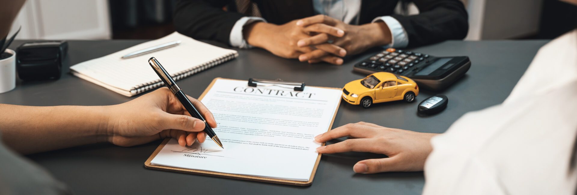 Two people sit at a desk with a contract, pen, notebook, calculator, toy car, and car keys; one person is signing the contract, carefully reviewing the details about vehicle loan interest before finalizing the agreement.
