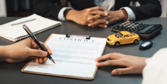Two people sit at a desk with a contract, pen, notebook, calculator, toy car, and car keys; one person is signing the contract, carefully reviewing the details about vehicle loan interest before finalizing the agreement.