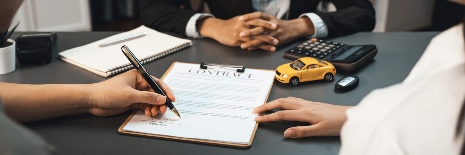 Two people sit at a desk with a contract, pen, notebook, calculator, toy car, and car keys; one person is signing the contract, carefully reviewing the details about vehicle loan interest before finalizing the agreement.