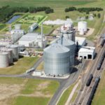 Aerial view of an ethanol production plant with large storage tanks, processing buildings, train tracks, and surrounding green fields.