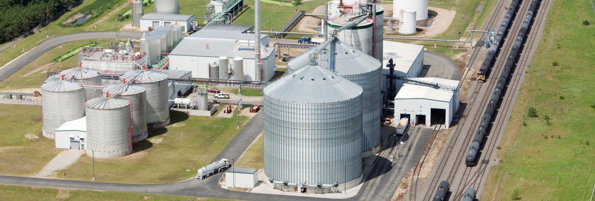 Aerial view of an ethanol production plant with large storage tanks, processing buildings, train tracks, and surrounding green fields.