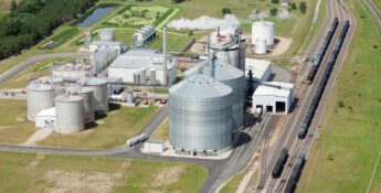 Aerial view of an ethanol production plant with large storage tanks, processing buildings, train tracks, and surrounding green fields.