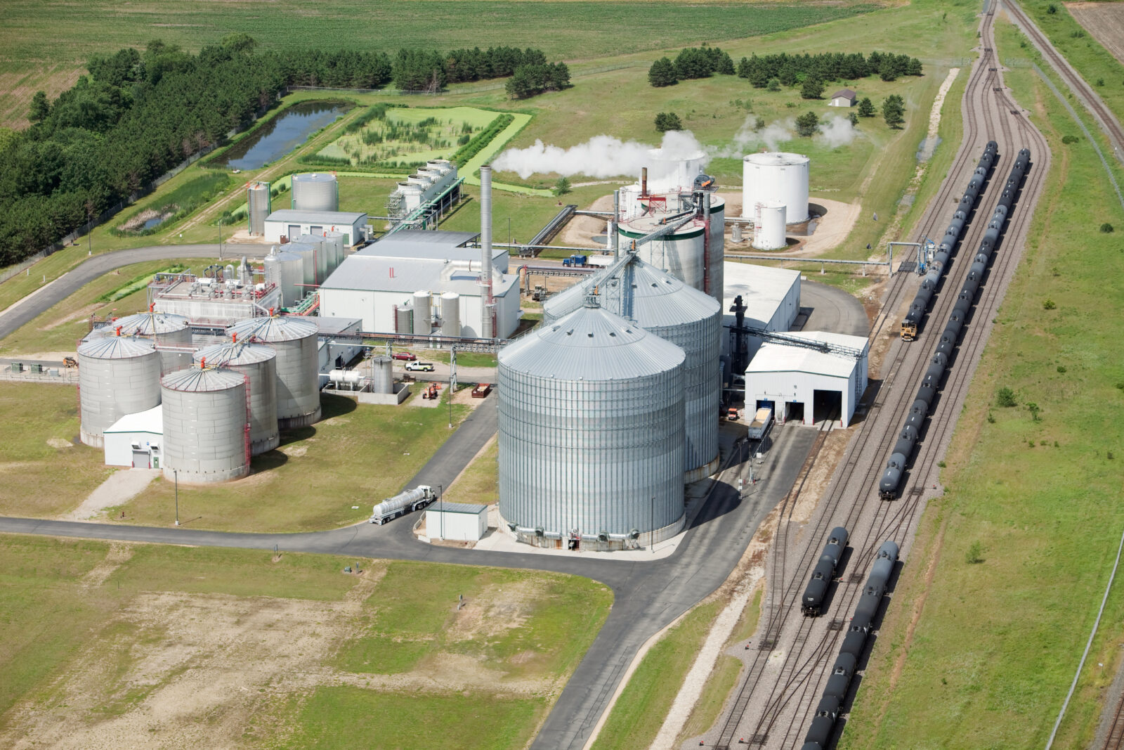 Aerial view of an ethanol production plant with large storage tanks, processing buildings, train tracks, and surrounding green fields.