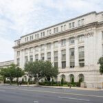A large neoclassical government building with columns and many windows, trees out front, and the Washington Monument visible in the background under a partly cloudy sky.