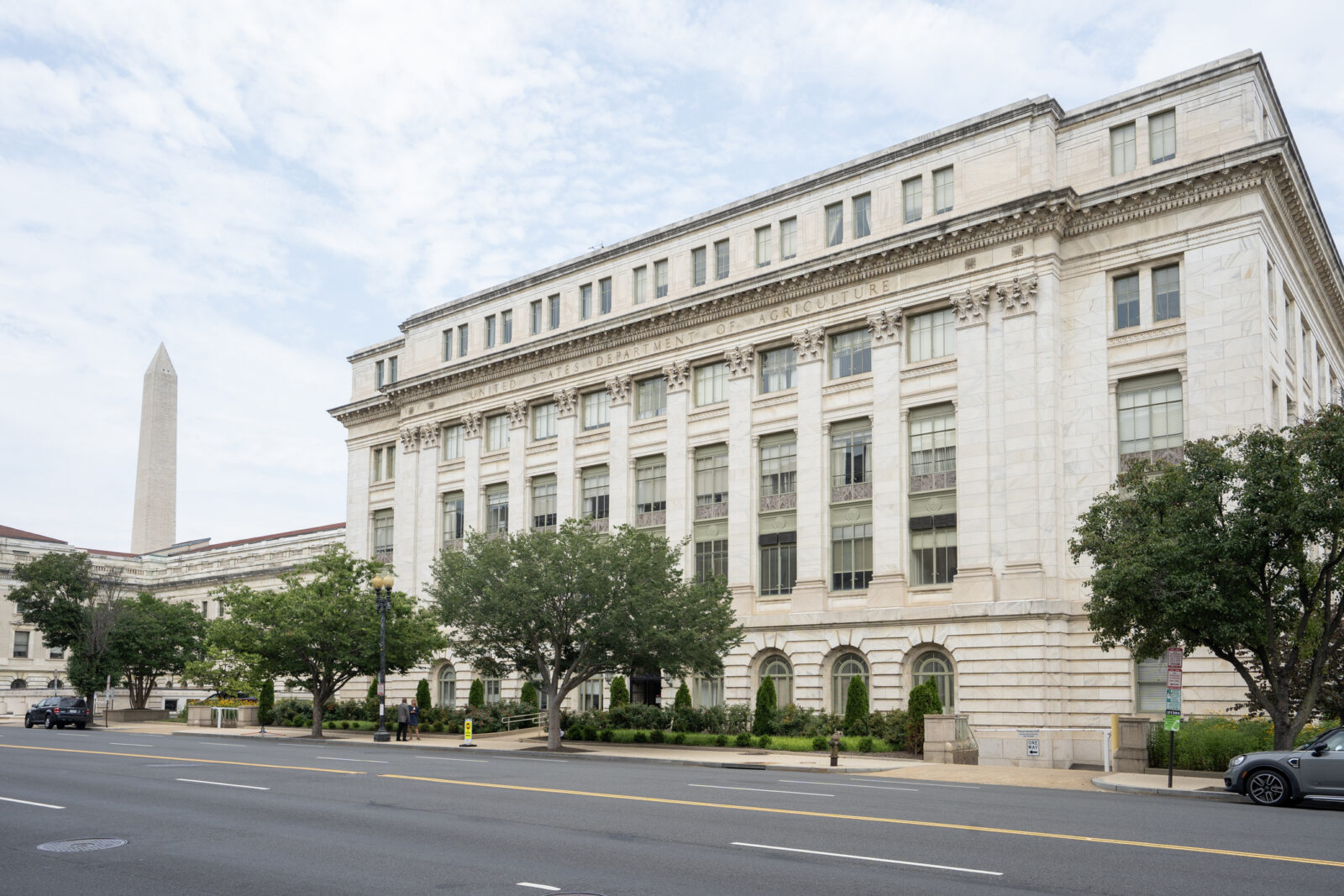A large neoclassical government building with columns and many windows, trees out front, and the Washington Monument visible in the background under a partly cloudy sky.