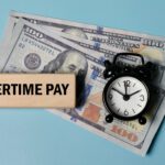 A wooden block labeled “Overtime Pay,” a small clock, and two one hundred dollar bills on a light blue background highlight the impact of the New Overtime Deduction Rules.