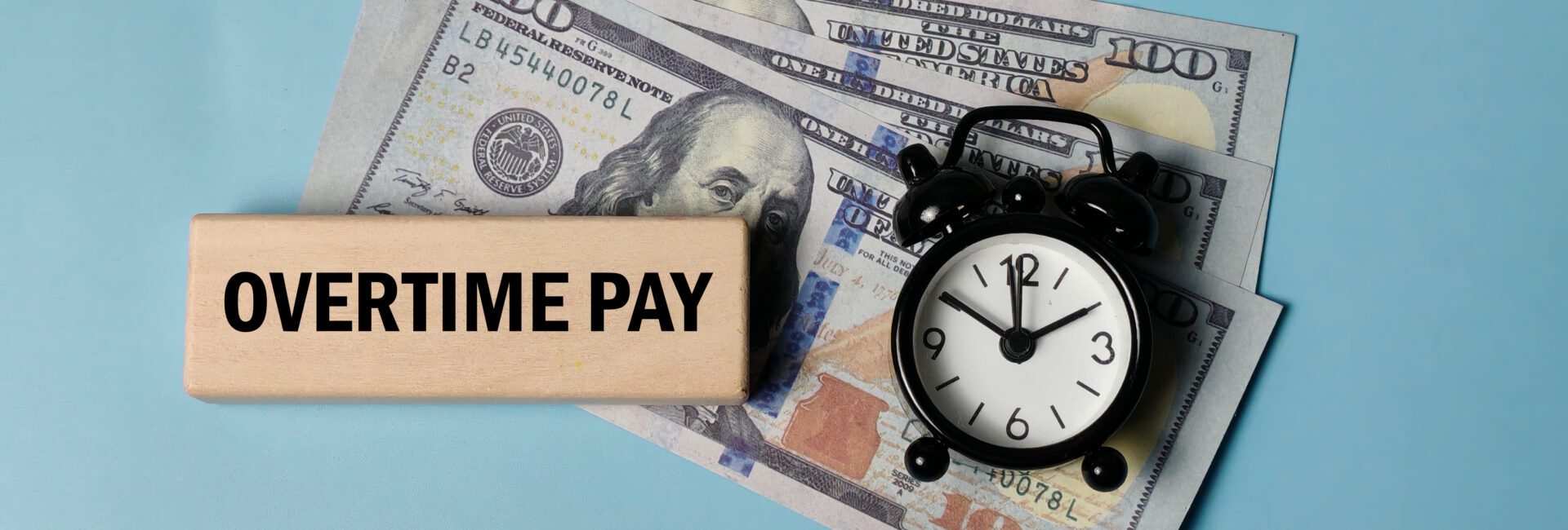 A wooden block labeled “Overtime Pay,” a small clock, and two one hundred dollar bills on a light blue background highlight the impact of the New Overtime Deduction Rules.
