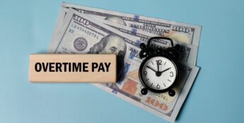 A wooden block labeled “Overtime Pay,” a small clock, and two one hundred dollar bills on a light blue background highlight the impact of the New Overtime Deduction Rules.