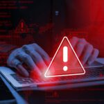 Hands typing on a laptop with a digital overlay of red warning icons and computer code.