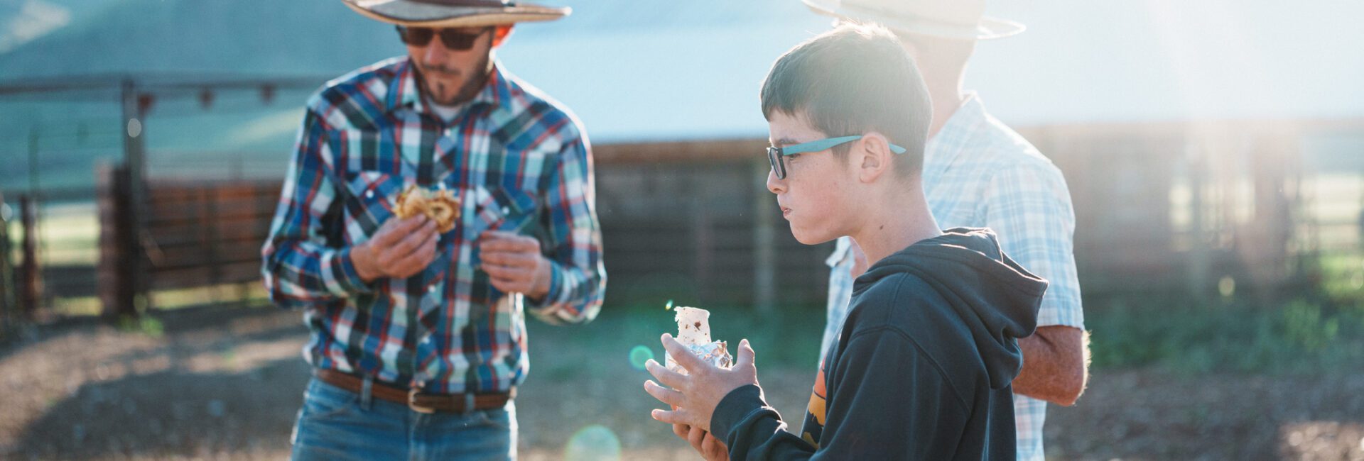 A young boy and two men in hats eating farm meals on a ranch.