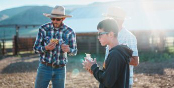 A young boy and two men in hats eating farm meals on a ranch.